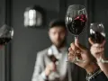 Wzniesione kieliszki z czerwonym winem, w tle mężczyzna w garniturze. Ciesz się chwilą z dobrym winem w kieliszku.