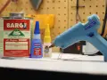 A hot glue gun, Barge cement, Loctite super glue, and Gorilla Super Glue are on a workbench. Does Gorilla Glue melt? This image shows various adhesives.