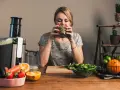 Kvinna dricker grön juice, omgiven av färsk frukt och grönsaker. Hon verkar njuta av sin hälsosamma dryck, kanske som en del av hur deffar man.