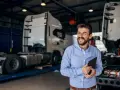 Uśmiechnięty mężczyzna z tabletem w ręku, w tle warsztat z ciężarówkami. Oferuje **usługę transportową ryczałt**.