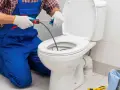 Instalator w niebieskim ogrodniczkach używa przepychacza do udrożnienia rury odpływowej WC.