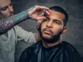 Barbero con tatuajes en el brazo recorta las cejas de un hombre con barba, creando un look buzz cut.