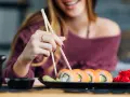 Uśmiechnięta kobieta w ciąży sięga po sushi pałeczkami. Na talerzu łosoś, awokado i imbir.