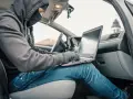 Haker w masce i kapturze próbuje obejść zabezpieczenia keyless go, używając laptopa w samochodzie.