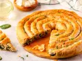 Złocisty borek ze szpinakiem, zwinięty w spiralę, kusi chrupiącym ciastem i aromatycznym nadzieniem. Obok kilka porcji i świeże liście szpinaku.