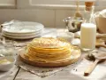 Une pile de crêpes dorées, prêtes pour une recette crêpe comme à la crêperie. Lait, crème et ustensiles à proximité.