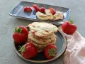 Ein Stapel Erdbeer-Pancakes mit frischen Erdbeeren auf einem Teller. Ein weiterer Teller im Hintergrund zeigt geschnittene Erdbeeren und Pancakes mit Erdnussbutter.