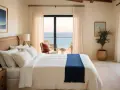 Acogedores dormitorios estilo mediterráneo con vistas al mar. Cama blanca con manta azul, sillón de mimbre y plantas.