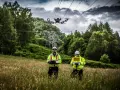 Two technicians in high-visibility jackets operate a power line drone in a grassy field, with a transmission tower and trees in the background.