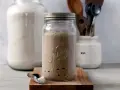 Blubbernde Sauerteig Kultur in einem Glas, bereit für das nächste Brot. Daneben ein Löffel mit Mehl und ein großer Behälter mit weiteren Zutaten.