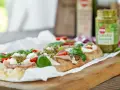 Długi placek z mozzarellą, pomidorkami i bazylią, polany zielonym pesto. Obok słoiczek z pesto.