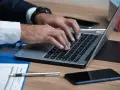 Ręce piszą na laptopie, obok telefon i długopis. Uważaj na oszustów, którzy podszywają się pod polskie firmy.