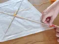 Mains fabriquant un cerf-volant simple en papier blanc et bâtons de bois, avec fil jaune.