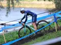 Cyclo cross racer in blue kit navigates a muddy course beside a lake, marked by blue tape.