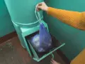 Ręka w żółtym swetrze wkłada worek ze śmieciami do metalowego zsypu w bloku.