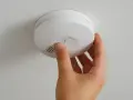 En hand trycker på testknappen på en vit brandlarm som sitter i taket. Hoppas att brandlarmet inte piper på riktigt.