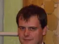 Tomasz Bartoś, zamyślony, w eleganckim stroju, na tle ozdobnej ściany.