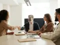 Uśmiechnięty mężczyzna ściska dłoń kandydatce, budując pozytywny employer branding. Przy stole siedzą też inne osoby.