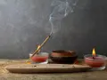 Un incienso humeante se posa sobre un soporte de arcilla, rodeado de velas rojas y un cuenco de madera. Perfectos inciensos para limpiar energías y crear un ambiente de paz.
