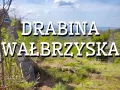 Widok na górskie krajobrazy z napisem "Drabina Wałbrzyska". Stary drewniany słup z niebieskim znakiem szlaku stoi na zboczu porośniętym trawą i młodymi drzewkami.