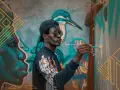 Un artiste au style d'art peinture, casquette et lunettes rondes, peint une fresque murale colorée représentant un visage et un oiseau.