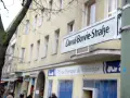 Straßenschild "David-Bowie-Straße" in Berlin. Ein Baum und ein Gebäude mit Geschäften im Hintergrund.