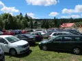 Tłum samochodów na parkingu przy Karłowie Szczeliniec. Słoneczny dzień, chmury na niebie, otoczenie lasu i budynków.