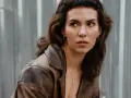 Nela Maciejewska w skórzanej kurtce, z burzą brązowych włosów i kolczykami w uszach, patrzy w bok na tle szarej, falistej ściany.