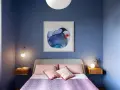 Camera da letto con pareti blu intenso e un quadro astratto. Cuscini rosa e coperta viola creano colori rilassanti.