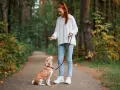 Rudowłosa dziewczyna uczy psa chodzić na smyczy w lesie. Pies siedzi, patrząc na nią z uwagą.
