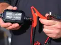 A person holds a NOCO Genius 2 trickle charger and its red clamp. This charger is designed to prevent damage, so no, a trickle charger can't ruin a battery when used correctly.