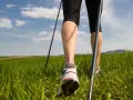 Gambe che praticano nordic walking tecnica su un prato verde sotto un cielo azzurro con nuvole bianche.