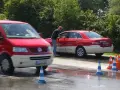 Mann reinigt Taxi. Ein roter Transporter fährt vorbei. Verkehrskegel markieren den Bereich. Möglicherweise eine Schulung der Berufsgenossenschaft Taxi.