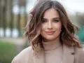 Kobieta z falowanymi włosami w stylu long bob, w beżowym płaszczu i golfie, uśmiecha się delikatnie.