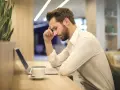 Mann mit Bart sitzt nach langer Krankheit wieder arbeiten ohne Wiedereingliederung am Laptop, die Stirn gerunzelt.