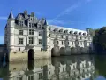 The stunning Chateau de Chenonceau, a white stone castle, gracefully spans the River Cher, its reflection shimmering on the water's surface under a clear blue sky.