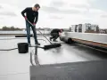 Dwóch mężczyzn kładzie papę na dachu. Jeden rozprowadza płynny dysperbent, drugi rozwija rolkę papy.