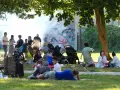 Familien genießen den Sommer auf einem Grillplatz in Berlin. Rauch steigt von einem Grill auf, während Kinder spielen und sich entspannen.