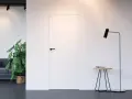 Białe drzwi ukryte w ścianie z czarną klamką, obok stojąca lampa i stolik.