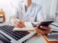 Lekarz w białym kitlu pisze w notatniku, trzymając telefon. Obok laptop, stetoskop i butelki.