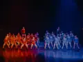Grupa tancerzy na scenie, podzielona na dwie grupy: jedna w pomarańczowych strojach, druga w niebieskich. Nauka tańca nowoczesnego w pełnym blasku.