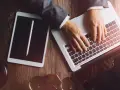 Dłonie na klawiaturze laptopa, obok tablet i młotek sędziowski. Profesjonalne narzędzia do pracy, być może związane z **taxat windykacja**.