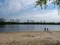 Zwei Kinder planschen im Wasser am Sandstrand des Dieksees in Lingen. Bäume säumen das Ufer unter einem bewölkten Himmel.