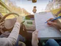 Egzamin na prawo jazdy. Kandydat trzyma kierownicę, a egzaminator zaznacza na liście kontrolnej różne kategorie prawa jazdy.