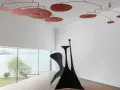 Un mobile Calder rouge flotte au-dessus d'une sculpture noire monumentale, dans une salle d'exposition lumineuse avec vue sur un lac.