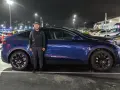 Mężczyzna stoi obok ciemnoniebieskiego Tesli Model Y na nocnym parkingu.