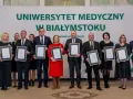 Grupa osób w garniturach i sukienkach pozuje do zdjęcia z dyplomami przed banerem Uniwersytetu Medycznego w Białymstoku.