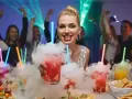 Dymiący drink przepis na imprezę. Kobieta z drinkiem w dymie, ludzie tańczą w tle.