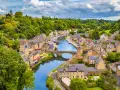 A picturesque view of Dinan, France, with its charming stone buildings lining a winding river, a stone bridge, and lush green hills under a cloudy sky.