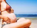Ciężarna kobieta na plaży, z kremem przeciwsłonecznym na brzuchu w kształcie słońca. Bezpieczne opalanie natryskowe w ciąży.